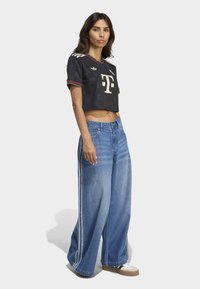 Donna che indossa un top sportivo nero a maniche corte con loghi, jeans larghi blu con strisce bianche laterali e sneaker bianche, in piedi su uno sfondo bianco.