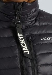 Gros plan sur une veste matelassée noire avec un col haut, une fermeture éclair proéminente et une languette en tissu noir portant l'inscription "JACK1T" en lettres blanches.