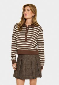 Femme portant un polo à rayures marron et beige à manches longues et une mini-jupe plissée marron avec un motif à carreaux subtil.