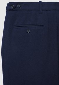 Gros plan sur un pantalon en laine bleu marine montrant la poche arrière avec fermeture à bouton et la ceinture avec deux boutons et passants de ceinture.