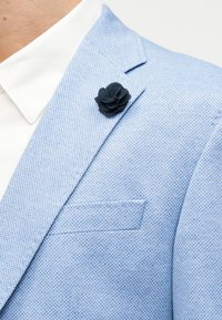 Blazer albastru deschis texturat cu o broșă floare neagră din material pe rever, peste gulerul unei cămăși albe.