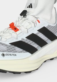 Λευκά και γκρι αθλητικά παπούτσια Adidas Gore-Tex με μαύρες ρίγες, κόκκινο βρόχο κορδονιού και υφασμένο πέλμα για εξωτερική χρήση.