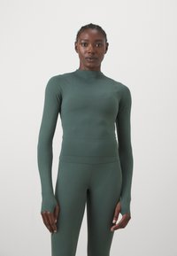 Langärmliges, eng anliegendes Sportoberteil aus tiefgrünem, glattem Stoff mit Daumenlöchern an den Bündchen. Passende hochgeschnittene Leggings.
