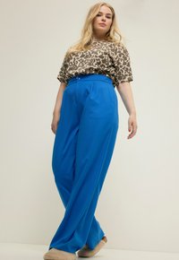 Een leopardprint top met korte mouwen gecombineerd met hoge, brede blauwe broeken. De broeken hebben een knoop bij de taille en een gladde textuur.