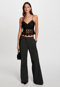 Zwarte kanten crop top met golvende randen, gecombineerd met hoge, op maat gemaakte zwarte broeken. Model houdt een kleine zwarte handtas vast.