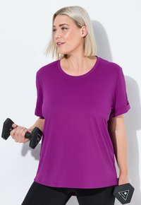 Kvinde med blond hår iført en lilla skjorte, der løfter sorte sekskantede håndvægte mod en hvid baggrund.