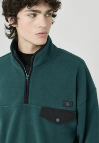 Pull en polaire vert foncé avec col zippé, poche noire et détails contrastants. Texture douce et coupe décontractée.