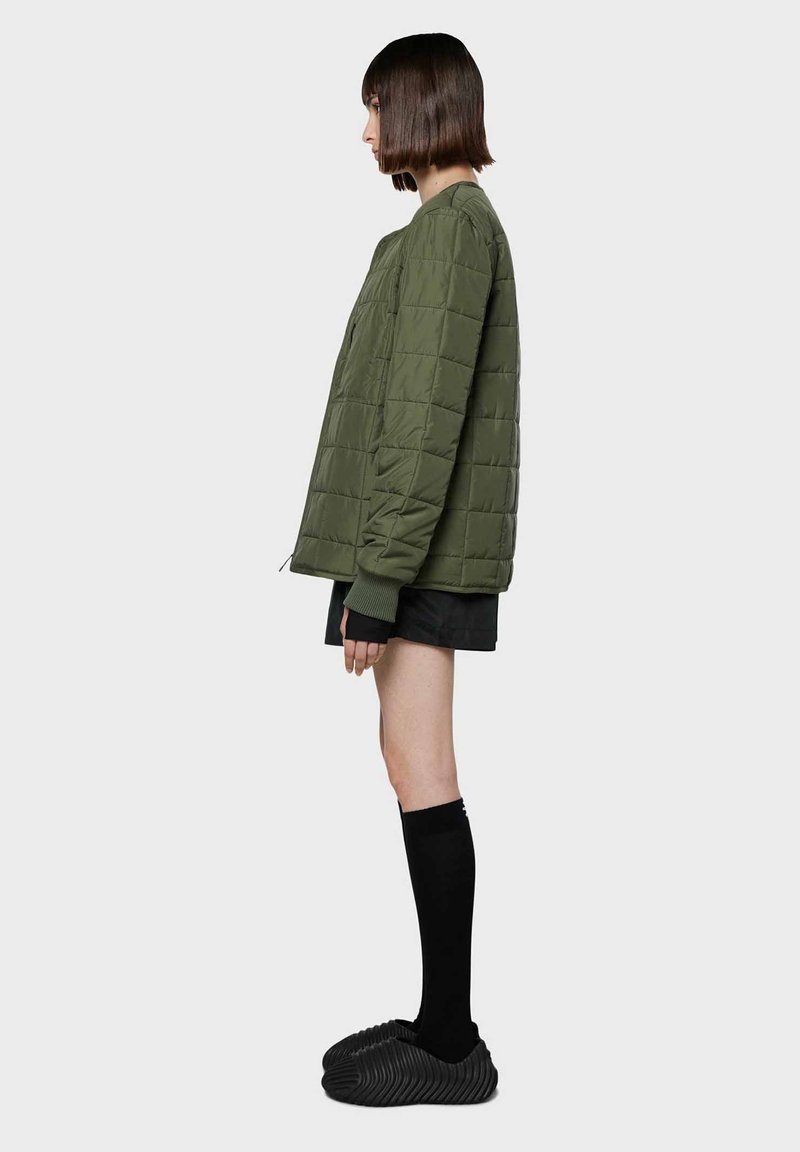 Giacca trapuntata verde oliva con vestibilità rilassata, maniche a coste e scollatura rotonda, abbinata a pantaloni corti neri e calze alte fino al ginocchio.