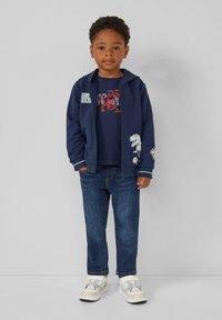 Marineblaues Zip-Top-Hoodie mit weißen Akzenten, blaues Grafik-T-Shirt und Jeans. Weiße und beige Sneakers. Enthält Dinosaurier-Motive auf der Kleidung.