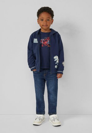 Marineblaues Zip-Top-Hoodie mit weißen Akzenten, blaues Grafik-T-Shirt und Jeans. Weiße und beige Sneakers. Enthält Dinosaurier-Motive auf der Kleidung.