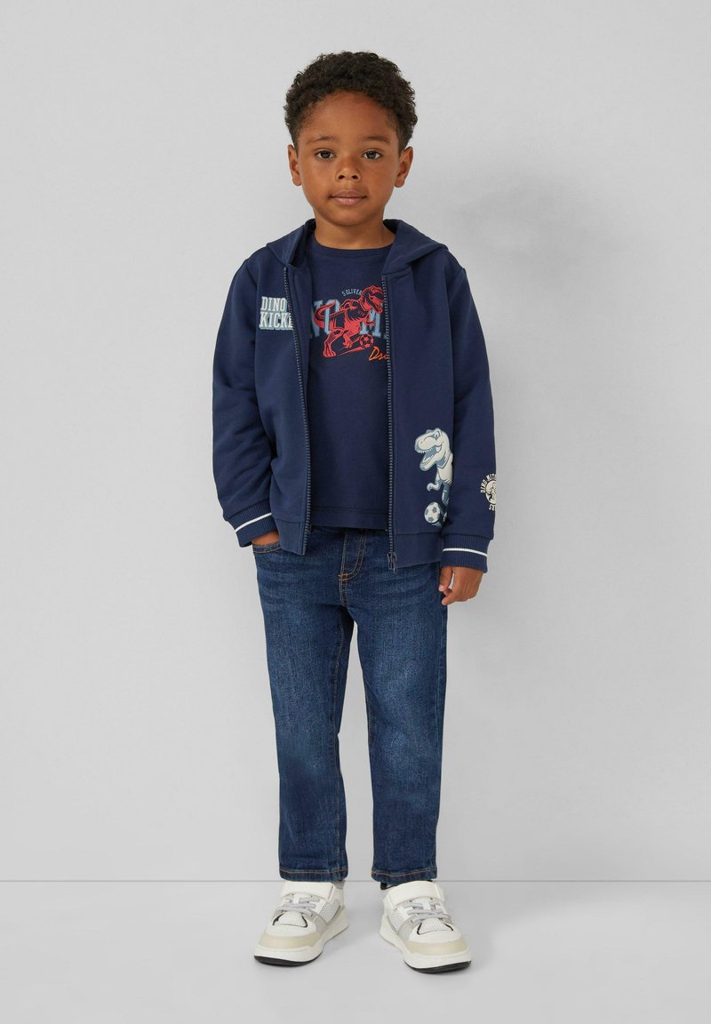 Marineblaues Zip-Top-Hoodie mit weißen Akzenten, blaues Grafik-T-Shirt und Jeans. Weiße und beige Sneakers. Enthält Dinosaurier-Motive auf der Kleidung.