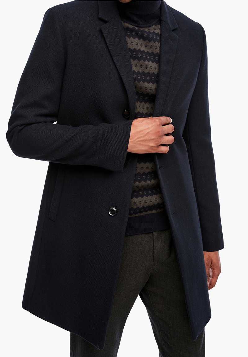 Marineblå frakke i uldblanding med hakkede revers, to knapper og tekstureret finish; lagdelt over mønstret sweater og båret med mørke bukser.