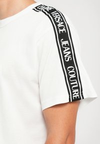 Balta trumpomis rankovėmis marškinėliai su juodu dryžiu ant peties ir rankovės, turintys balta spalva užrašytą "Versace Jeans Couture" tekstą.