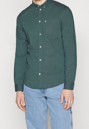 Homme portant une chemise à boutons vert foncé avec une poche sur la poitrine et un jean bleu clair, avec une bague argentée à la main gauche.