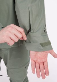 Zeleni jakni materijal s munjevitom džepom na rukavu, podesivim manžetama s čičak trakom i glatkom teksturom. Ruka prikazana za mjeru.