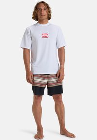 Biała koszulka z krótkim rękawem typu rash guard z czerwonym logo oraz kolorowe kąpielówki w paski w odcieniach niebieskiego, pomarańczowego i beżowego, z ciemniejszym niebieskim dolnym fragmentem.