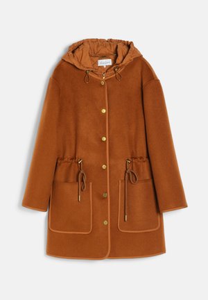 Jeune femme portant un manteau cape marron par-dessus une veste matelassée, un pantalon vert et de longues bottes marron, debout devant un fond uni.
