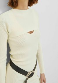 Kvinna i en krämfärgad ribbad stickad klänning med ett urtag vid bröstet och ett brett svart bälte med en silverfärgad spänne.