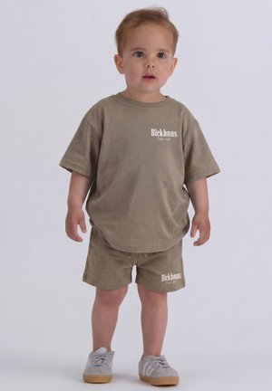 Peuter die staat, gekleed in een bijpassend bruin T-shirt en korte broek met witte tekst en lichtgrijze sneakers, op een effen lichte achtergrond.