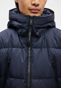 Tengerészkék pufi kabát magas gallérral, cipzáras záródással és dupla húzózsinórral. Átlósan steppelt mintázat jellemzi, valamint két cipzáras oldalsó zseb van rajta.