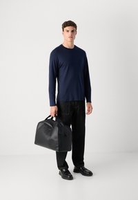 Man wearing navy long-sleeve shirt, black pants, and black shoes holding large black leather tote bag against white background.