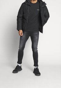 Černá péřová bunda s kapucí, černé tričko, tmavé džíny a černé tenisky. Obsahuje zipové zapínání a boční kapsy.