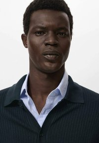 Giovane uomo con pelle scura e capelli ricci corti, che indossa una camicia azzurro chiaro sotto un cardigan scuro a coste, in piedi davanti a uno sfondo bianco puro.