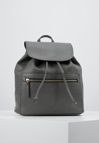 Sac à dos en cuir gris avec fermeture à rabat, cordon de serrage, poche avant zippée et fermetures éclair dorées. Texture lisse et design minimaliste.