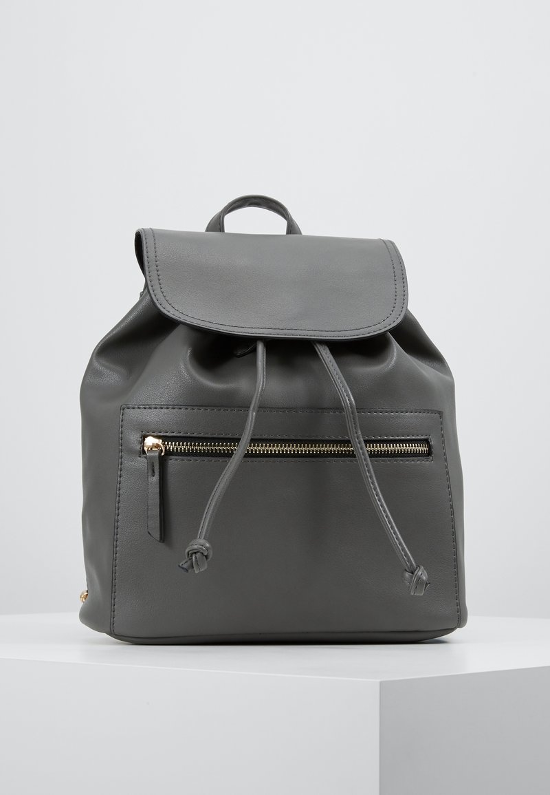 Sac à dos en cuir gris avec fermeture à rabat, cordon de serrage, poche avant zippée et fermetures éclair dorées. Texture lisse et design minimaliste.