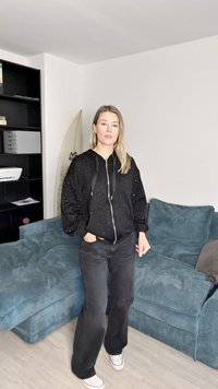 Kvinne med langt blondt hår iført svart hettegenser og bukser, som står foran en blå sofa og en svart bokhylle med et surfebrett bak.