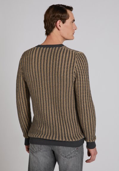 Hombre con un suéter de punto marrón y negro con un patrón texturizado, visto desde atrás, combinado con jeans grises sobre un fondo liso.