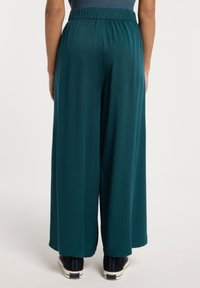 Pantaloni larghi color teal con elastico in vita, caratterizzati da una texture morbida e un leggero lucido, abbinati a scarpe scure su uno sfondo neutro.