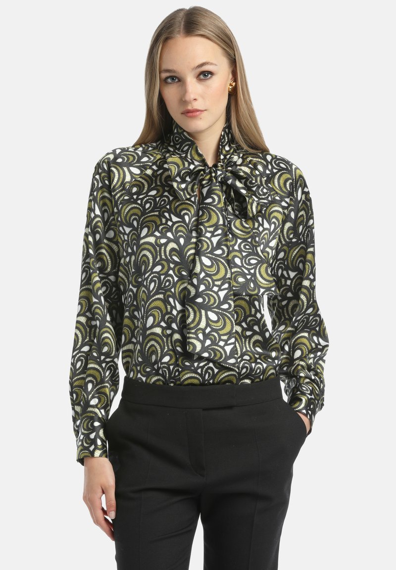 Blusa bianca e nera con un motivo paisley verde, caratterizzata da maniche lunghe, un collo a fiocco e una texture satinata liscia. Indossata con pantaloni neri.