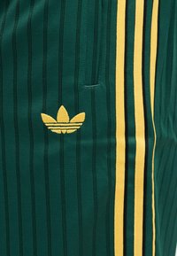 Πράσινο ύφασμα με λεπτές μαύρες κάθετες ρίγες, κίτρινο λογότυπο Adidas trefoil και κίτρινες και μαύρες κάθετες πλευρικές ρίγες.