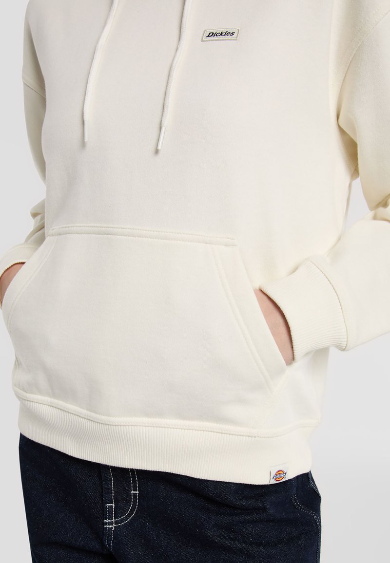 Sudadera color crema con bolsillo canguro, puños y dobladillo acanalados; cuenta con un pequeño parche del logo de Dickies en el pecho. Material suave y casual.