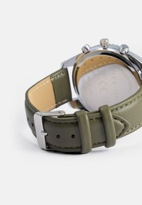 Zaļš ādas pulksteņa siksniņa ar sudraba metāla sprādzi un šuvēm. Apaļš nerūsējošā tērauda korpuss ar gludu aizmuguri.