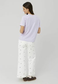 Un t-shirt lilas à col rond associé à un pantalon large blanc orné de broderies florales. Des chaussures marron et des chaussettes mi-mollet complètent la tenue.