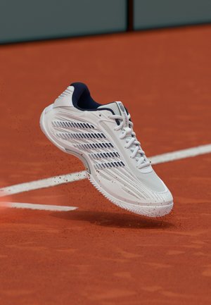 Bela športna obutev s črnimi črtami dviguje rdeč prah na teniškem igrišču blizu bele mejne črte.