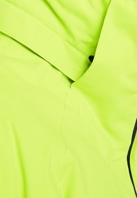 Tissu jaune-vert vif avec des coutures visibles, une sangle pliée et un petit bouton noir partiellement caché dans une couture.