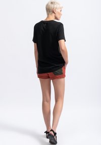 Zwarte T-shirt met ronde hals, gecombineerd met roestkleurige shorts met een donkergroene zak. Model staat zijwaarts en toont de details van de outfit.