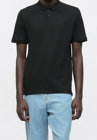 Czarna koszulka polo z klasycznym kołnierzykiem, krótkimi rękawami i subtelnym logo. Połączona z jasnoniebieskimi dżinsami.