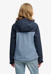 Osoba o blond włosach do ramion ubrana w dwukolorową niebieską kurtkę z kapturem i jasnoniebieskie dżinsy, pokazana od tyłu.