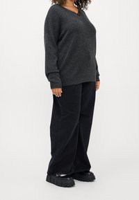 Kvinna klädd i en mörkgrå tröja, svarta vida byxor och kraftiga svarta skor som står mot en vit bakgrund.