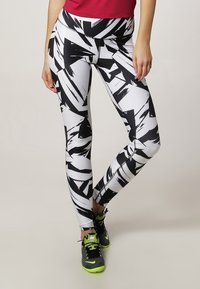 Mujer con leggings blancos con patrones geométricos abstractos en negro, zapatillas deportivas negras y verde neón, y una blusa roja, de pie con las piernas cruzadas.