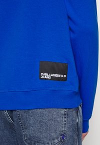 Plava dukserica s rebrastim manžetama i porubom, sa crnom tkaninom oznakom s natpisom "KARL LAGERFELD JEANS." U kombinaciji s iznošenim trapericama.