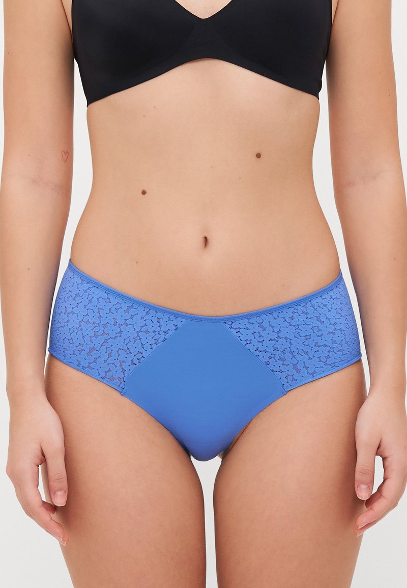Culotte hipster bleue avec un motif en dentelle texturée sur les côtés et un devant en tissu lisse et unis, présentant un design à taille mi-haute.