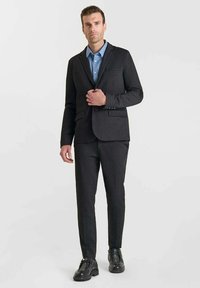 Completo grigio scuro con vestibilità slim, caratterizzato da un rever a punte, chiusura con un solo bottone e pantaloni davanti piatti abbinati a una camicia azzurro chiaro e scarpe nere.