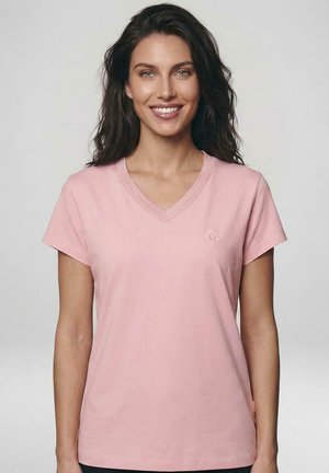 Lachende vrouw met lang donker haar die een lichtroze T-shirt met V-hals en korte mouwen draagt tegen een effen lichte achtergrond.