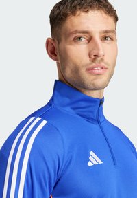 Kék sportos cipzáros dzseki fehér csíkokkal a vállakon és egy adidas logóval a mellkason; sima anyag, testhezálló fazon.