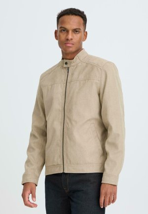 Man draagt een beige jack met ritssluiting en opstaande kraag, en een donkere spijkerbroek, staande tegen een effen lichte achtergrond.